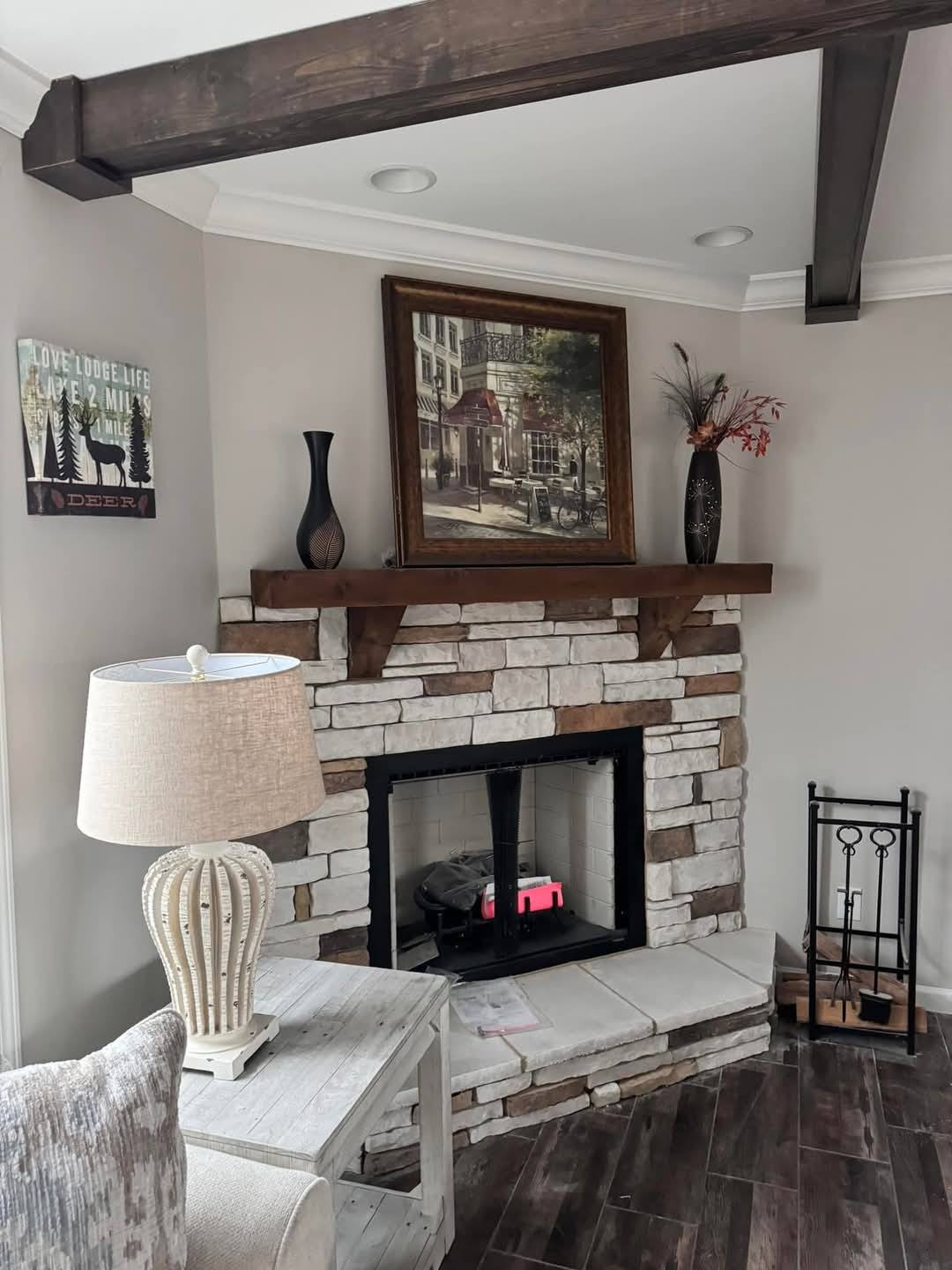 Cozy living room with a brick fireplace, wooden mantel, and artwork above. A rustic lamp and table sit nearby. Warm, inviting atmosphere.