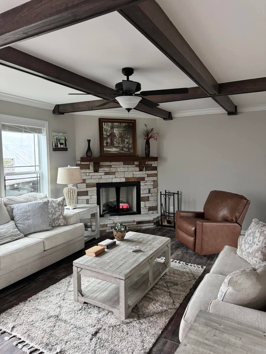 Cozy living room with a stone fireplace, wooden ceiling beams, and neutral tones. Features a beige couch, brown leather chair, and rustic decor.