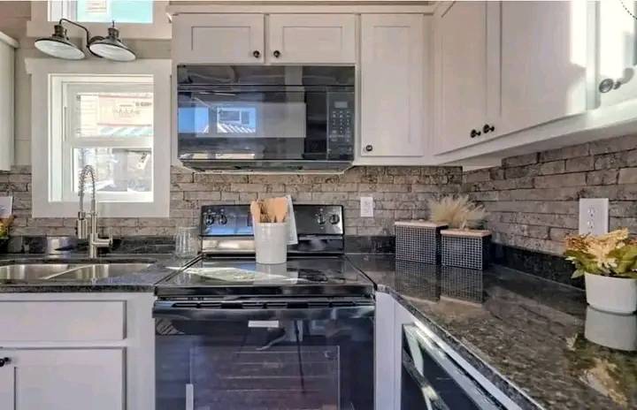 Modern kitchen with white cabinets, a stainless steel stove, microwave, and double sink. Brick backsplash and granite countertops add elegance.