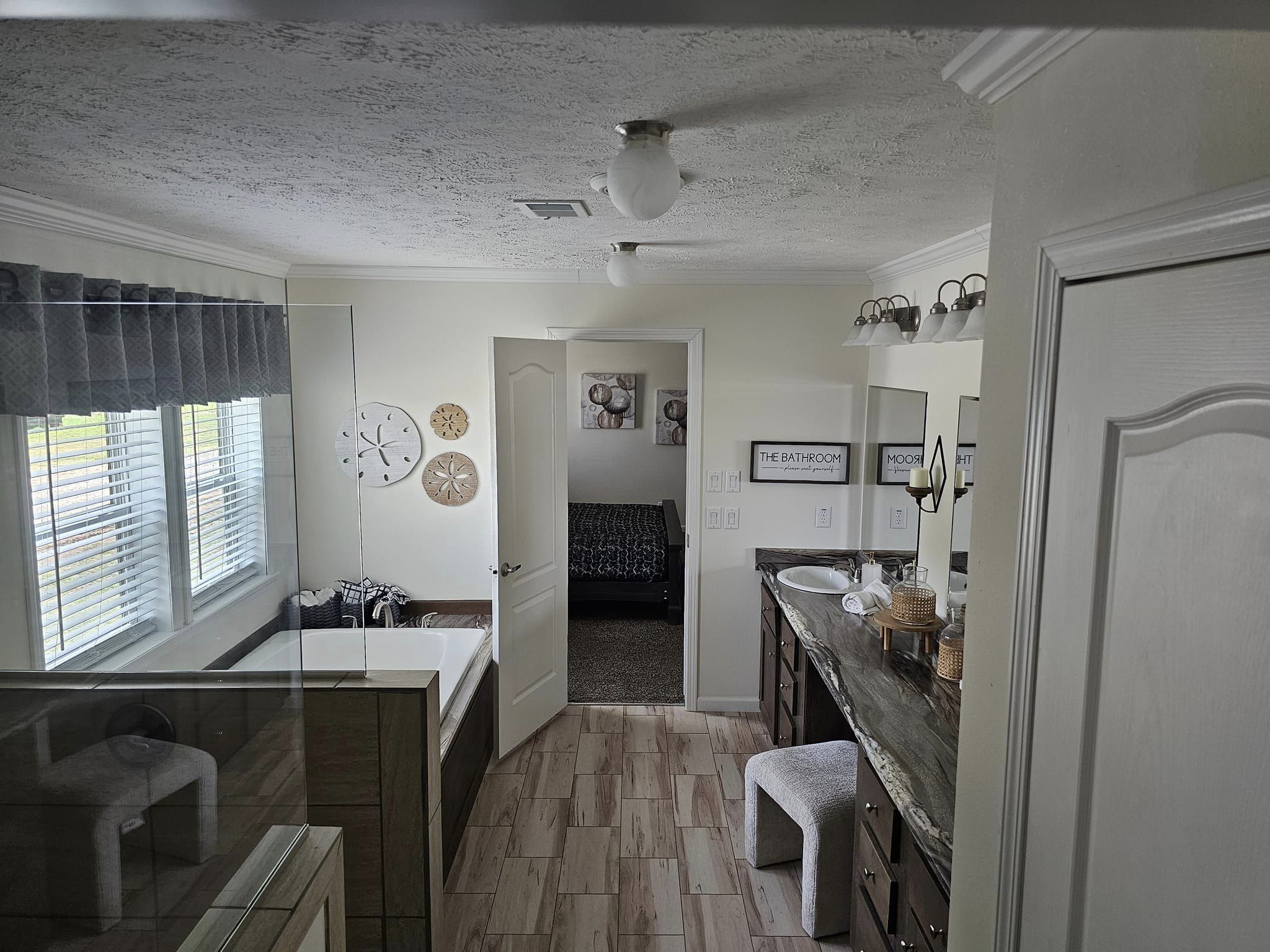 Bright bathroom with natural light from large windows on the left. Features a bathtub, double vanity with mirrors, stool, and art on walls. Cozy ambiance.