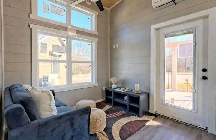 Cozy modern living room with large windows, blue sofa, beige pouf, and decorative pillows. Sunlit, warm tones create an inviting atmosphere.