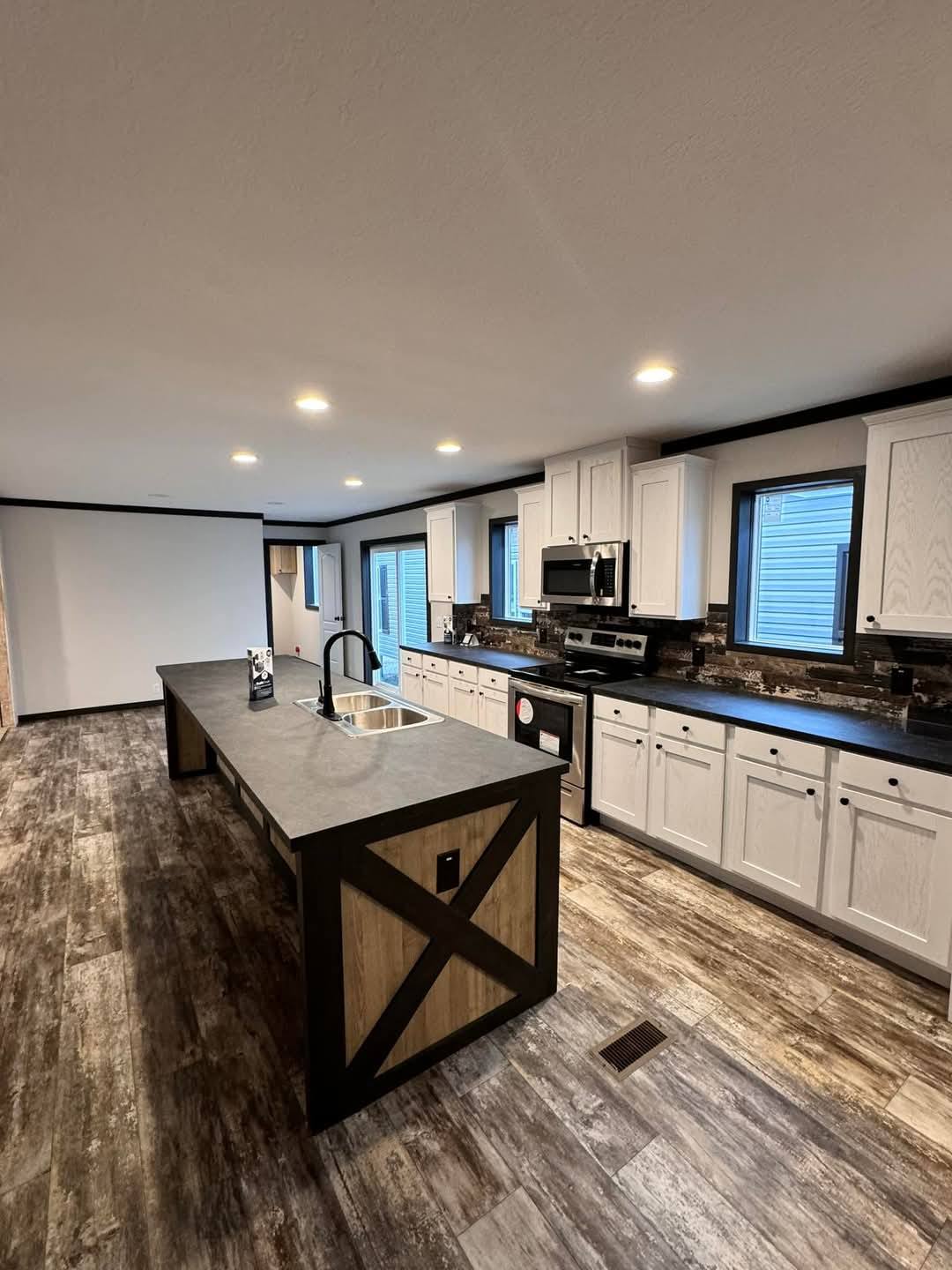 Modern kitchen with rustic elements, featuring a long island with a sink, white cabinets, stainless-steel appliances, and wood-look flooring. Warm lighting creates a cozy ambiance.