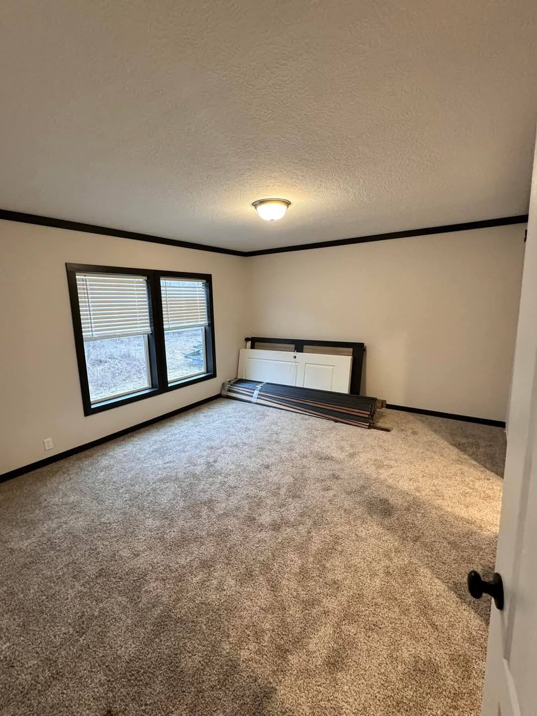 Empty room with soft beige walls and carpeting, illuminated by a ceiling light. Two windows with blinds overlook a bare outdoor scene. Disassembled door and wooden planks rest against a wall, conveying a sense of ongoing renovation or preparation.