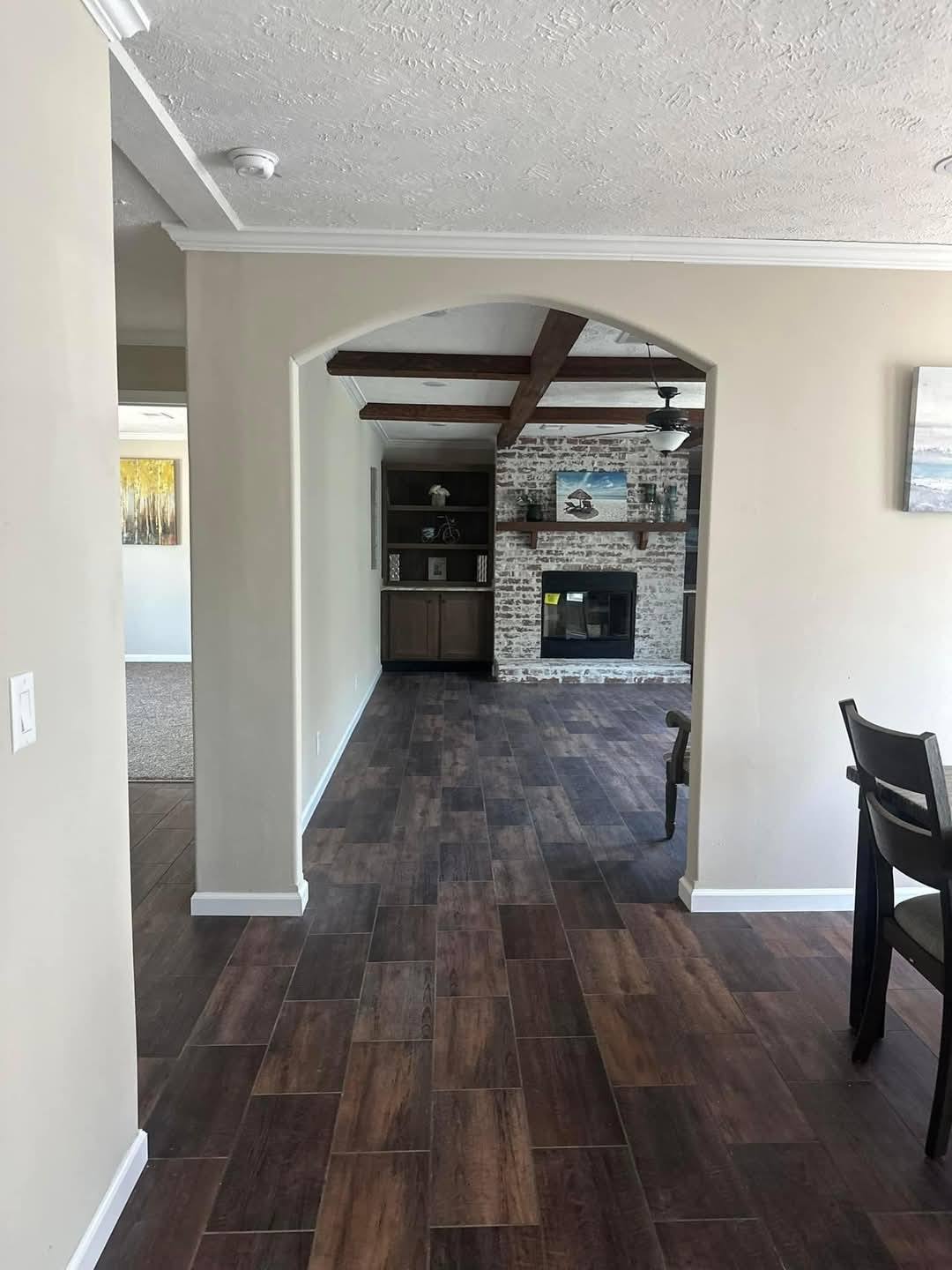 Spacious room with dark wood floors, a white brick fireplace under wooden ceiling beams, and neutral walls. A chair is partially visible on the right.
