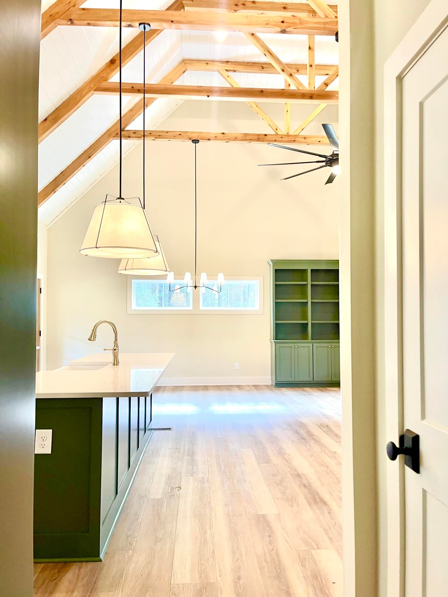 A bright, modern kitchen with light wood floors and vaulted ceiling with wooden beams. Features include a green island, pendant lights, and built-in cabinetry.