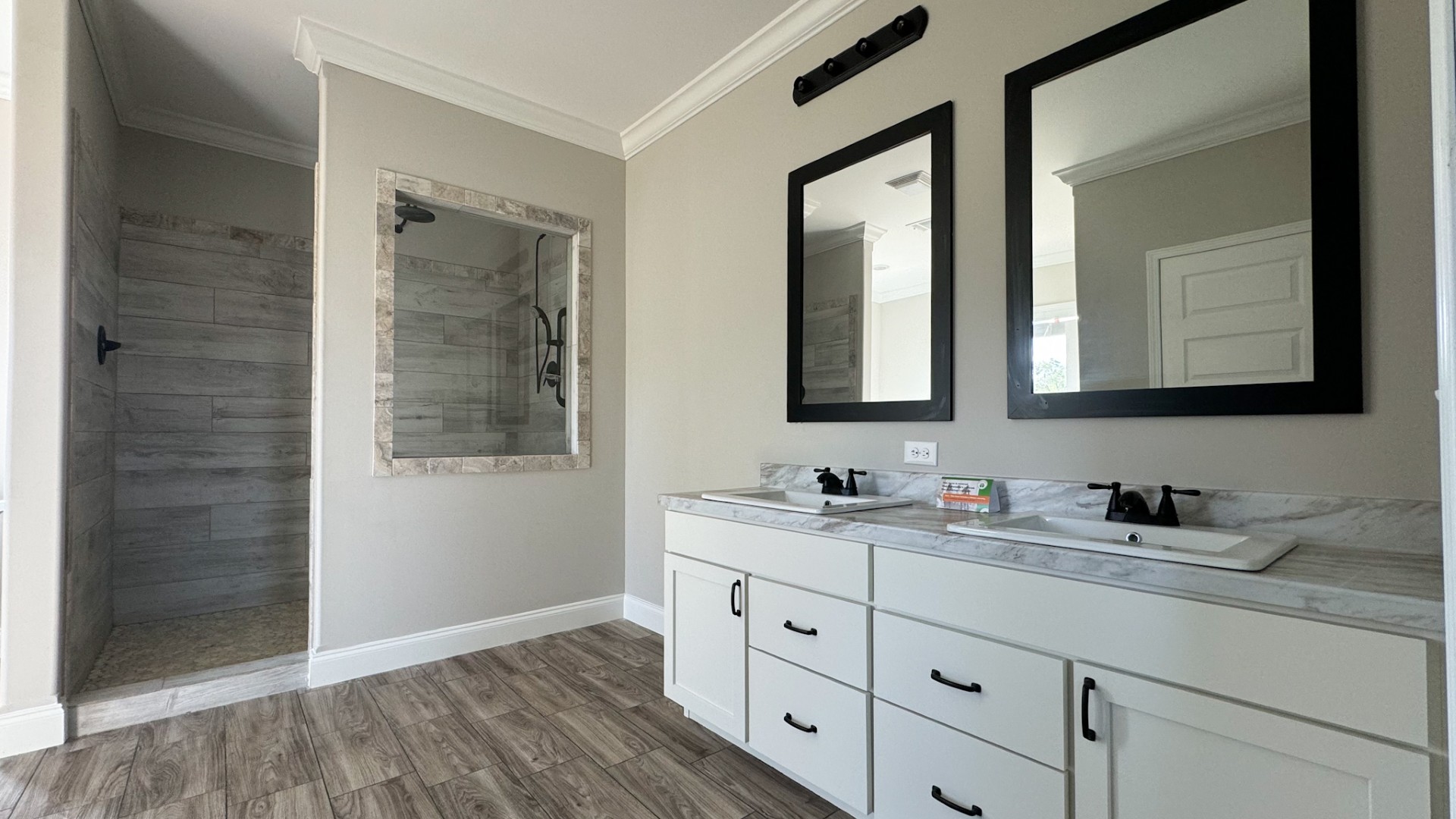 Modern bathroom with twin mirrors above a double sink vanity, white cabinetry, and wood-style flooring. A walk-in shower is visible on the left.