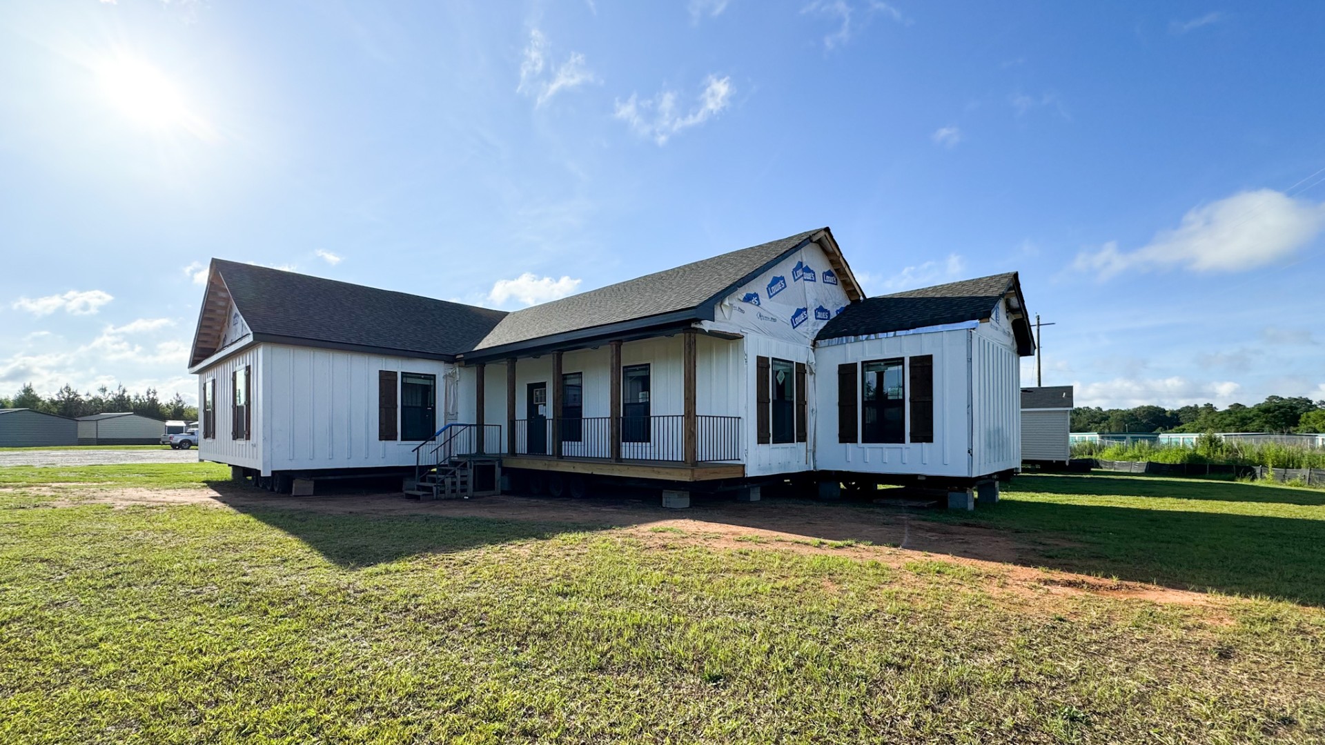 Alt text: "A newly constructed house stands on a grassy field under a bright blue sky. The home features white siding, dark roofing, and visible construction wrap, creating a fresh and optimistic atmosphere."