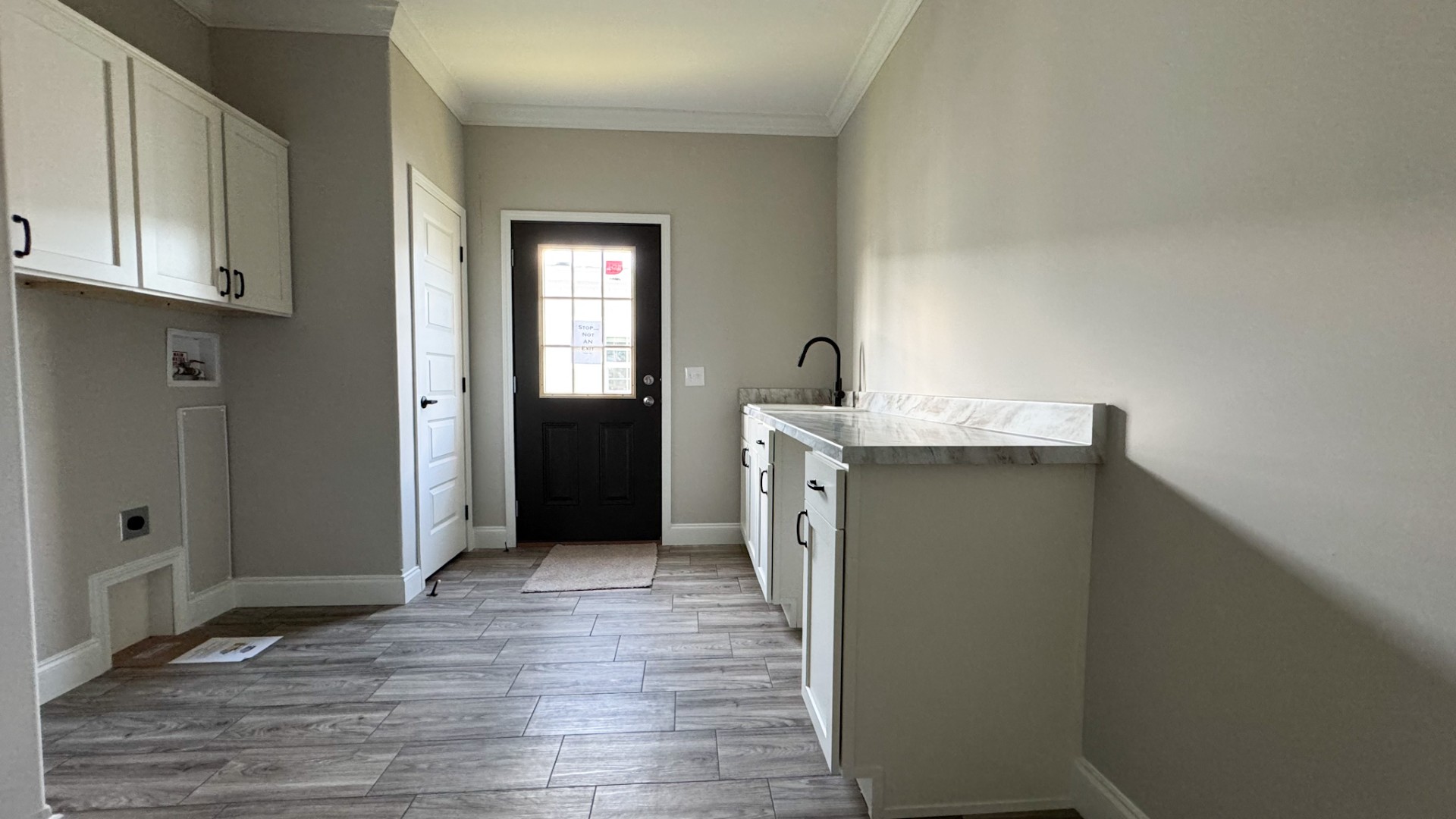 A modern laundry room with light gray walls and wood-patterned tile flooring. Features include white cabinets, a marble countertop, and a black door.