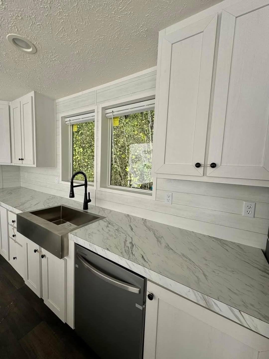 Modern kitchen featuring white cabinets, marble countertops, and a stainless steel sink with a black faucet. Large windows offer natural light.