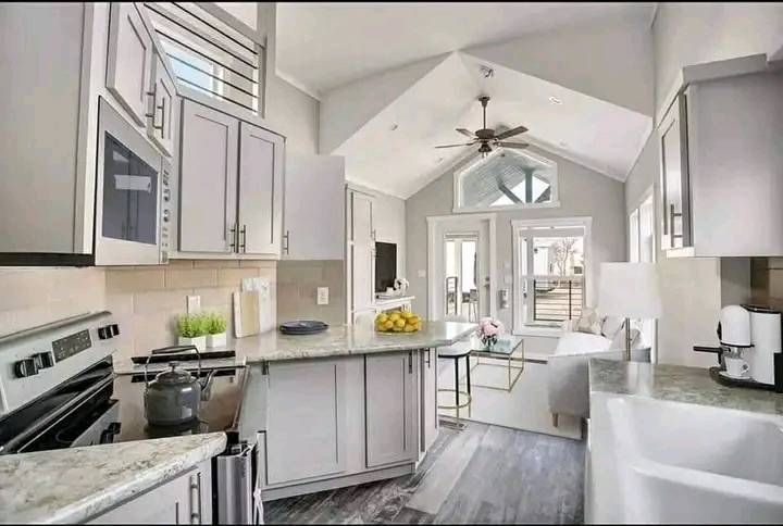 Modern kitchen and living area with vaulted ceilings. Light gray cabinets, fruit bowl on counter, and large windows fill space with natural light. Cozy, inviting atmosphere.