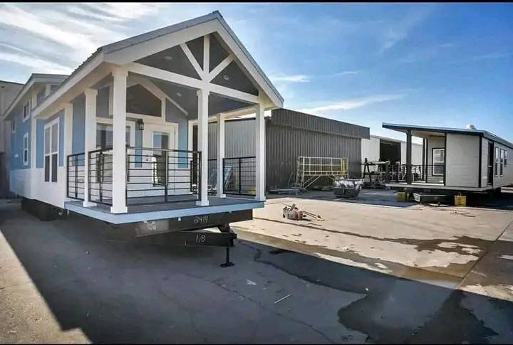 Alt text: "A light blue tiny house on a trailer with a white porch sits in an industrial lot under a clear blue sky. Another tiny house is visible in the background."