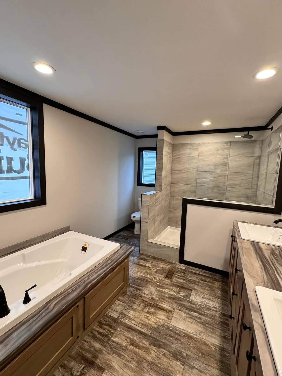 Modern bathroom with a spacious layout featuring a large bathtub on the left, a glass-enclosed shower in the background, and a sleek double vanity on the right. The room is well-lit with recessed ceiling lights and has a calm, neutral tone.
