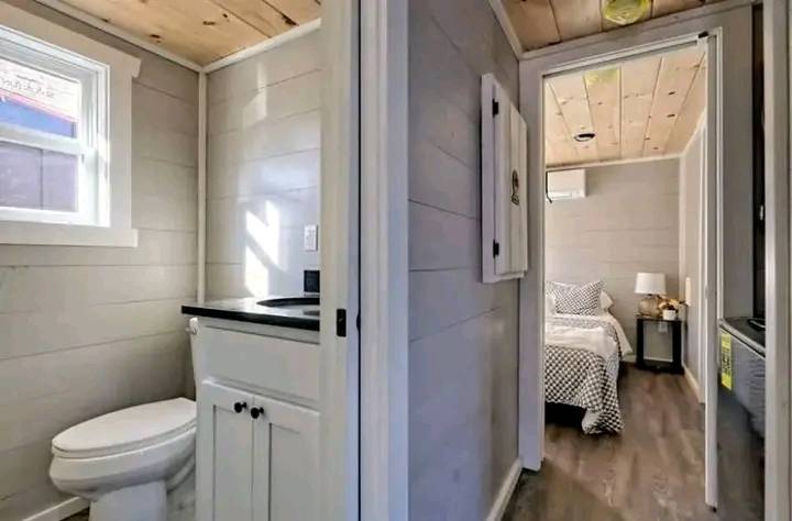 Compact bathroom and bedroom with wooden walls and ceiling. Bathroom has a toilet and sink; bedroom features a bed with patterned linens. Cozy and neat.