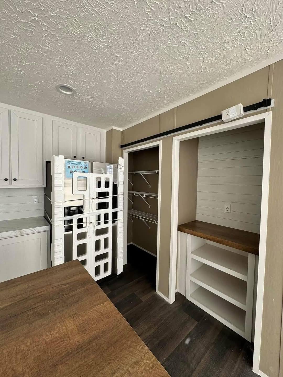 A small, modern kitchen with white cabinets, dark wood countertop, and an unpacked oven wrapped in white packaging. Light brown walls and a textured ceiling.