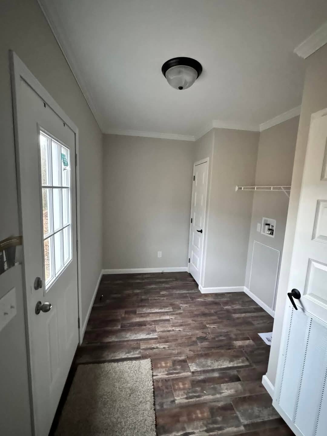 Narrow mudroom with wood-patterned floor, beige walls, and a white door with panes. Wall-mounted shelf, ceiling light, and a small rug add minimalistic charm.