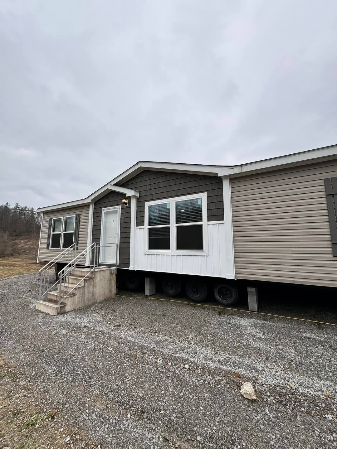 A mobile home with brown and white siding sits on a gravel lot under an overcast sky. Stairs lead to the front door, and trees are visible in the background.