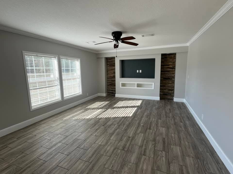 Spacious, empty living room with light gray walls and wood-look tile flooring. Features large windows, a ceiling fan, and a recessed wall with wooden accents.