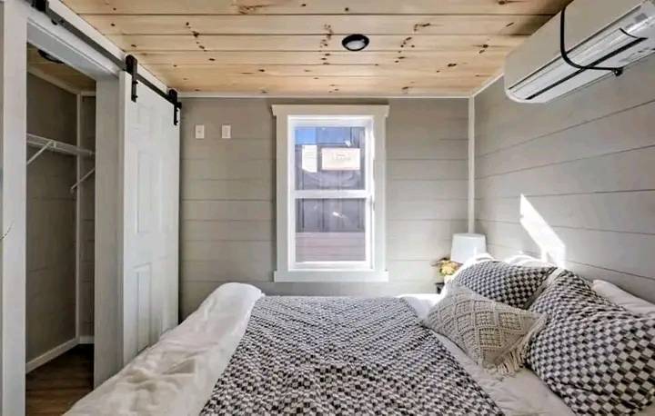 Cozy bedroom featuring a wooden ceiling, gray shiplap walls, a small window, and a bed with patterned pillows. An open closet and air conditioner add functionality.