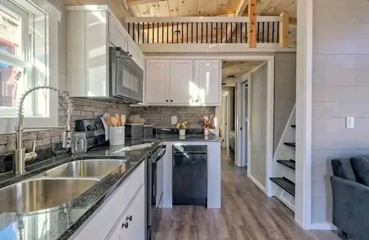 Modern tiny house kitchen with white cabinets, granite countertops, and stainless steel appliances. A loft with wooden railing is visible above. Cozy and bright.