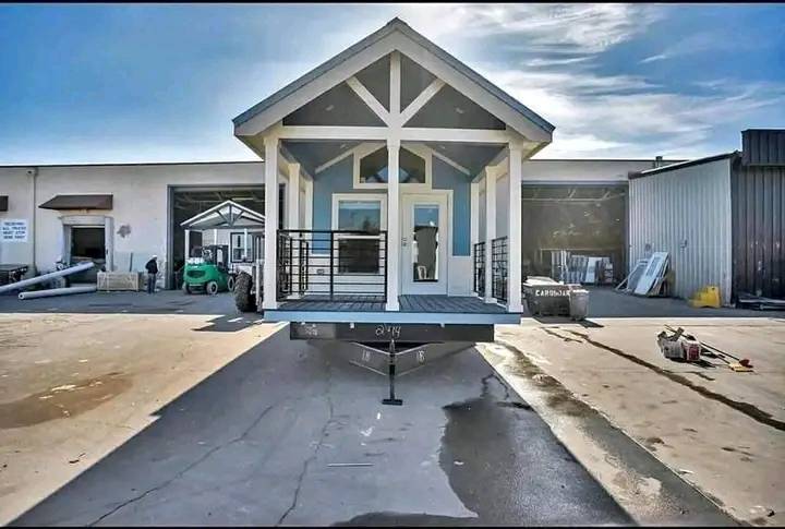 A modern tiny house on wheels with a blue and white exterior is parked outside a warehouse. The scene is sunny and industrial, suggesting construction.