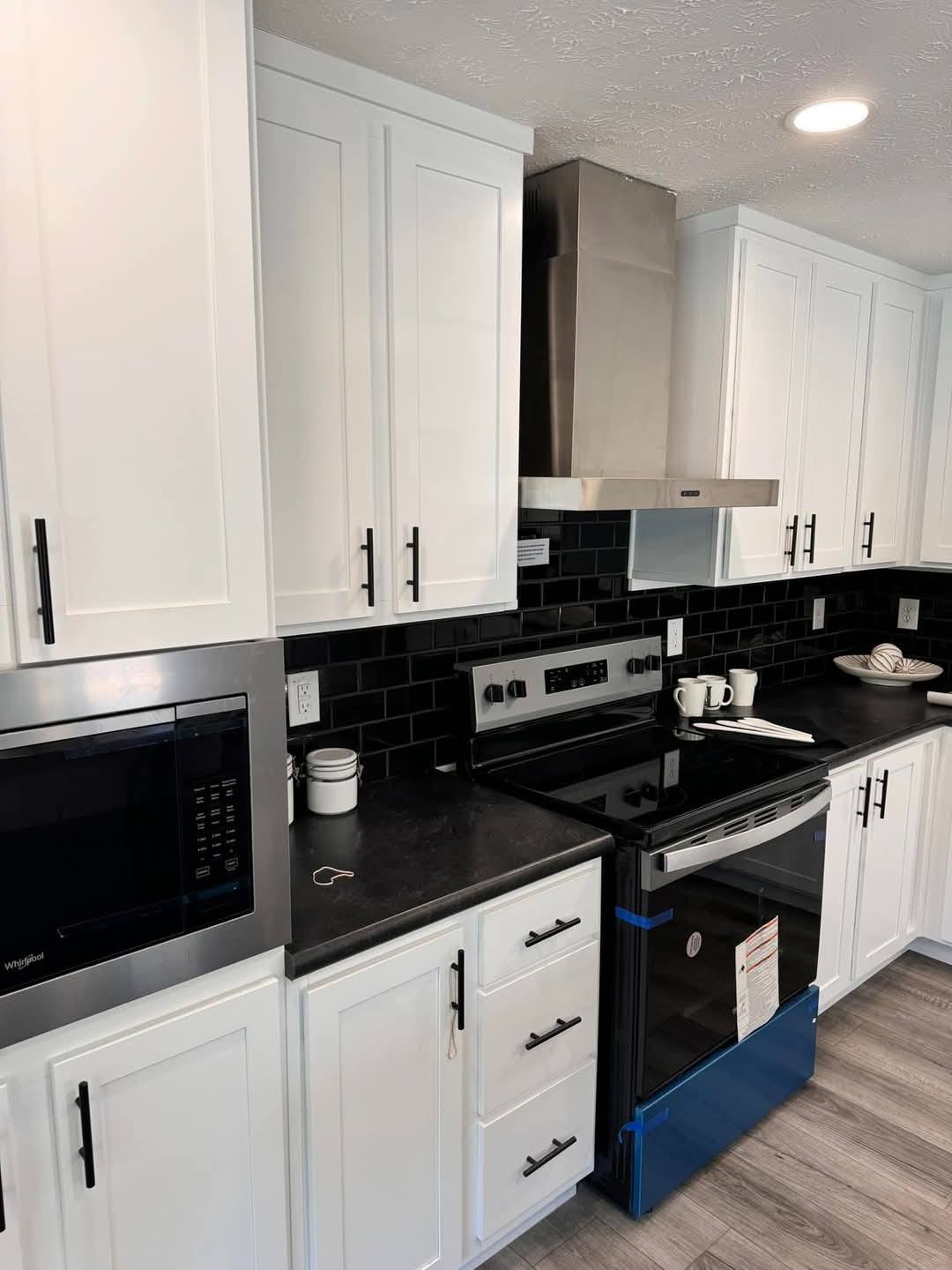 Modern kitchen with white cabinets, black countertops, and a stainless steel vent hood. Black tile backsplash adds contrast. Warm, inviting atmosphere.