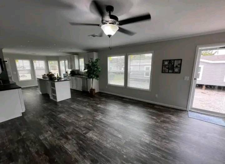 Spacious living area with dark wood floors, large windows, and a ceiling fan. White kitchen with island in the background; a calm, airy ambiance.