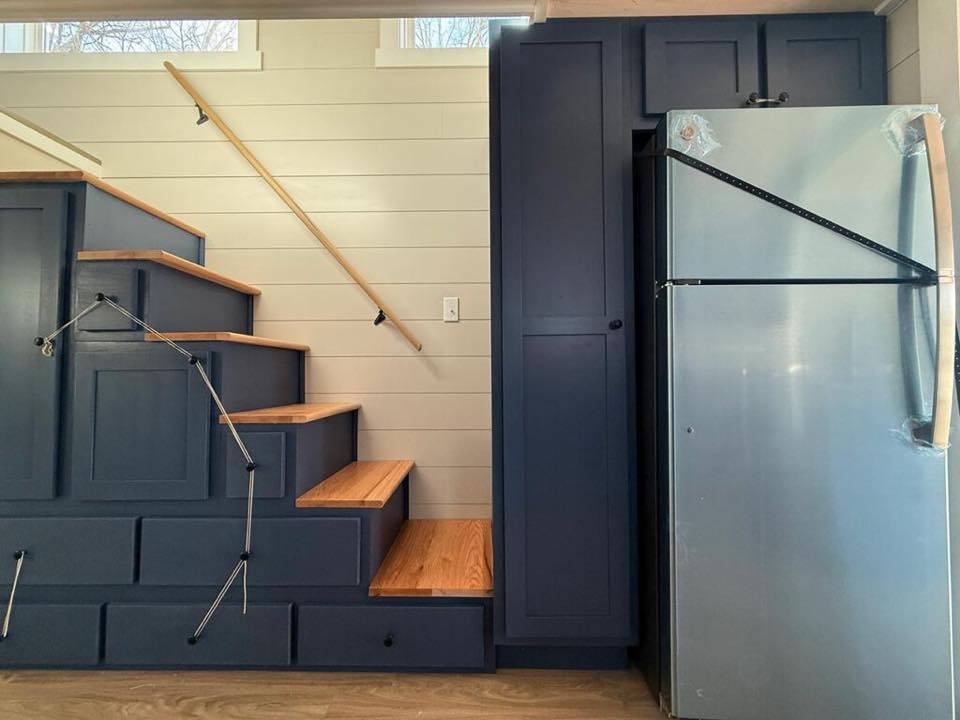 A modern kitchen with a sleek design features a metal staircase with wooden steps on the left, contrasting with dark blue cabinetry and a silver refrigerator.
