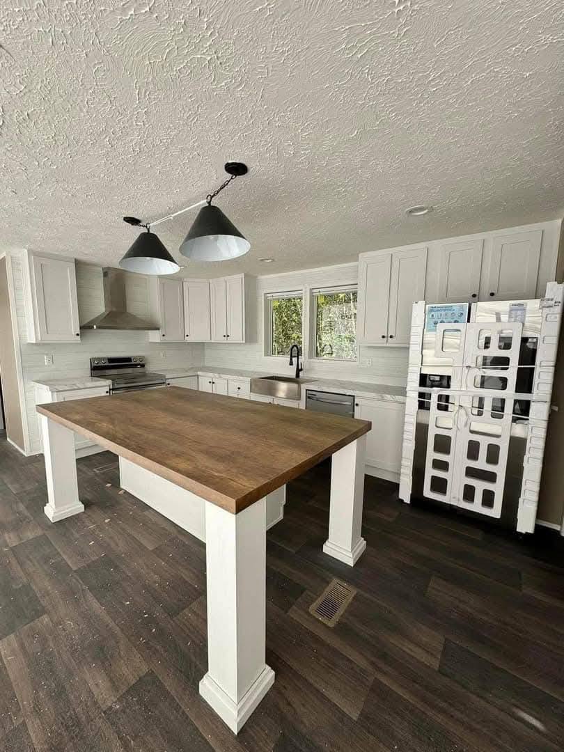 Modern kitchen with white cabinets, dark wood floor, and a central island with a wooden countertop. Stainless steel appliances and large windows add brightness.
