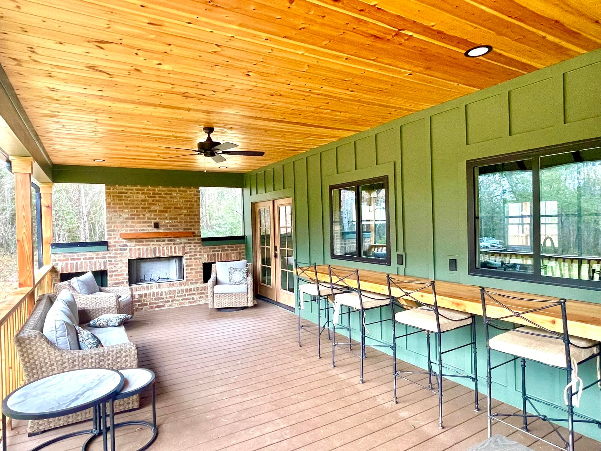 Covered porch with wooden ceiling, green walls, and brick fireplace. Features wicker furniture with cushions and a bar area with high chairs. Cozy and inviting.