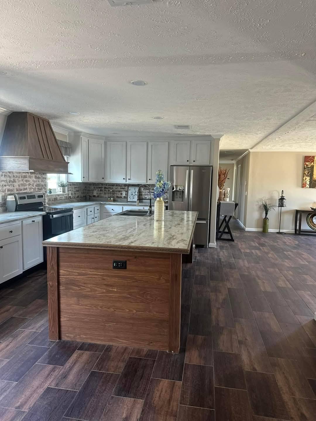 Bright kitchen with wooden flooring, centered by a marble-topped island. White cabinets, stainless steel appliances, and a rustic hood enhance elegance.