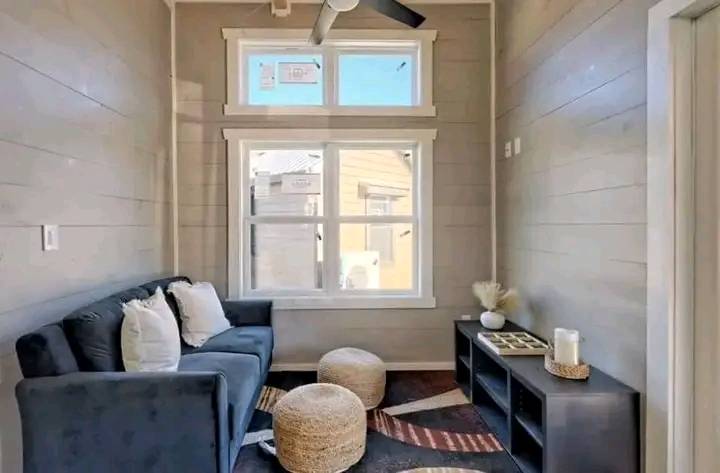 A cozy, narrow living room with light gray walls features a blue couch adorned with white pillows, two woven poufs, a patterned rug, a black shelf with decor, and a large window letting in sunlight. The tone is warm and inviting.
