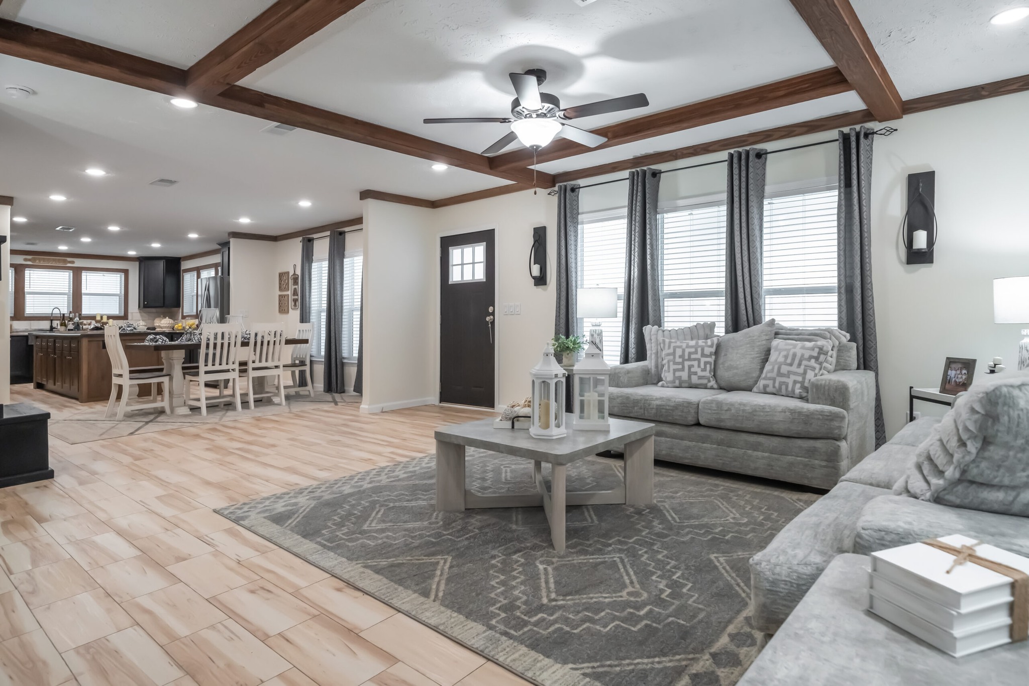 Spacious living room with gray sofas, a patterned rug, and wooden ceiling beams. Open kitchen and dining area in the background, natural light from windows. Cozy and inviting ambiance.