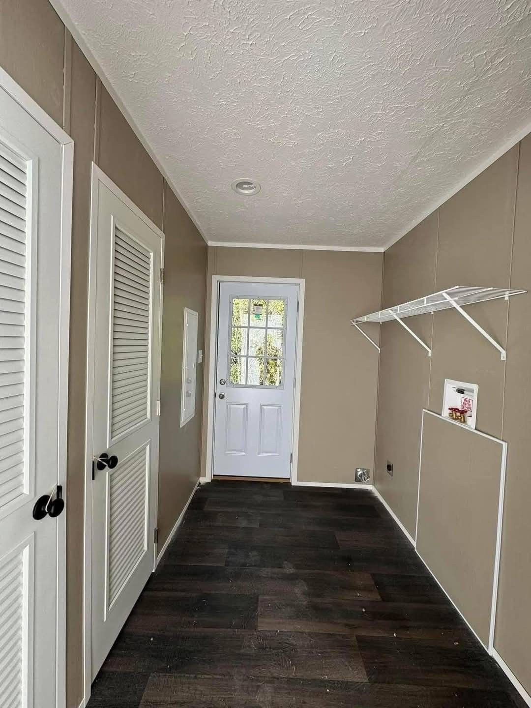 Narrow mudroom with beige walls, white louvered doors on the left, a glass-panel door at the end, dark wood flooring, and a wire shelf on the right.