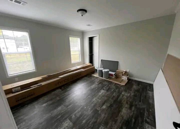Empty room with dark wood-style flooring, light gray walls, and two windows. Boxes and items are stacked near a door, indicating a move or renovation.