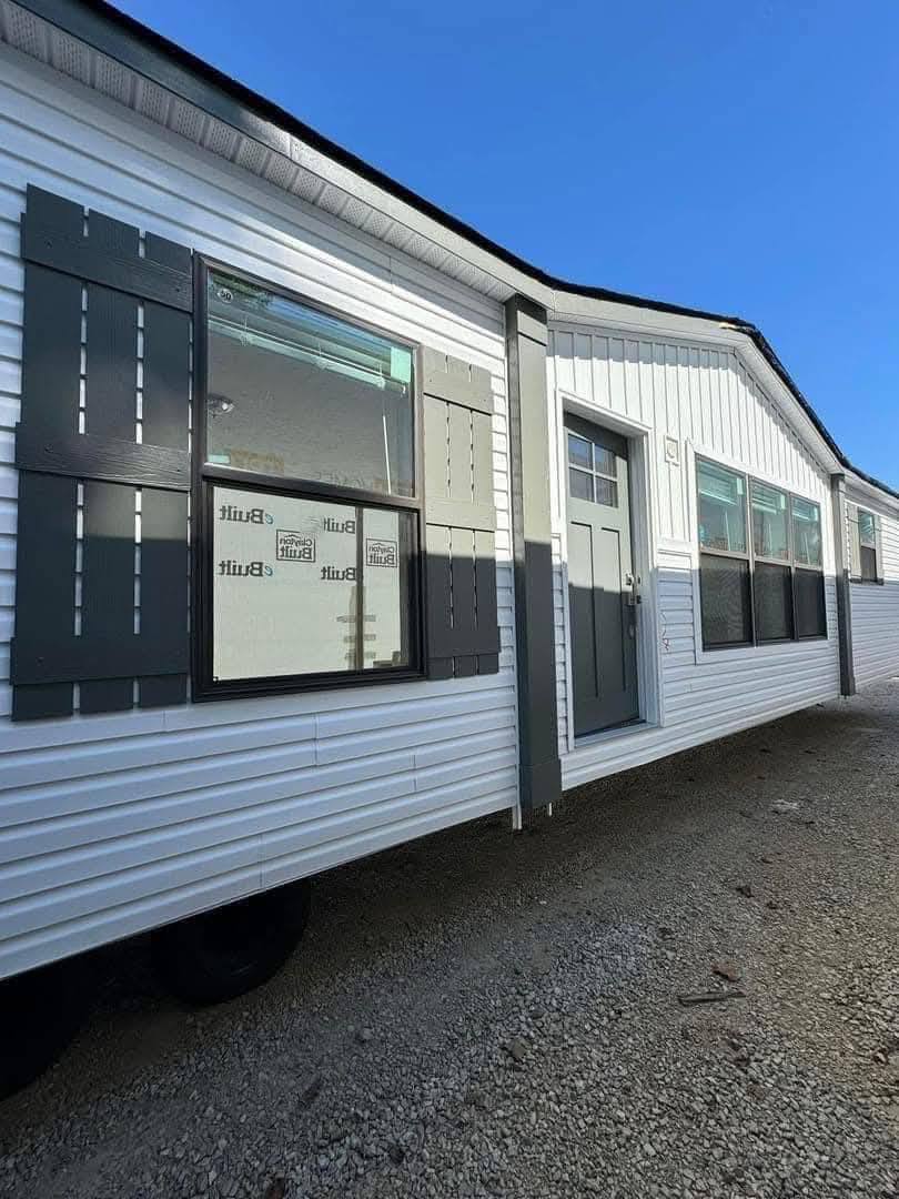 A white mobile home with gray shutters and door sits on a gravel lot under a clear blue sky. Sunlight casts shadows, creating a serene, inviting atmosphere.