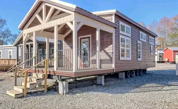 A modern tiny house on wheels with a covered porch and multiple windows, set on a gravel lot. The tone is inviting and cozy.