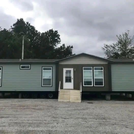 A mobile home with gray siding and white-trimmed windows sits on a gravel lot. Overcast sky above and trees in the background create a serene atmosphere.