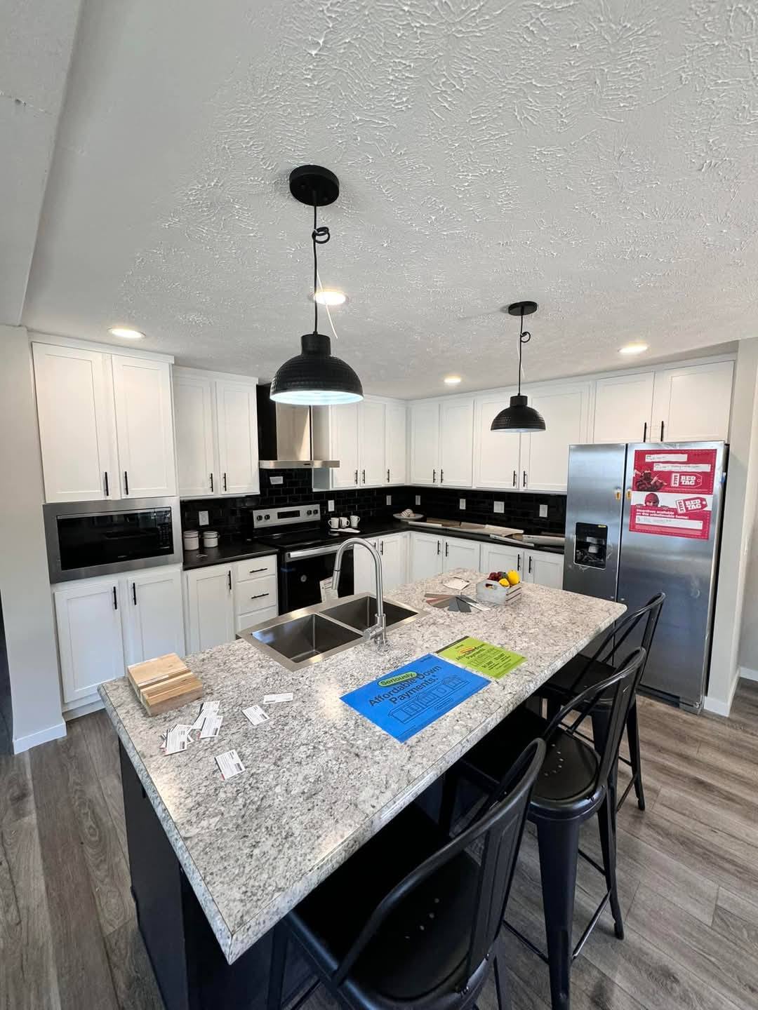 Modern kitchen with white cabinets, black accents, and stainless steel appliances. Highlights include a granite island with bar stools and pendant lighting. Warm, inviting atmosphere.