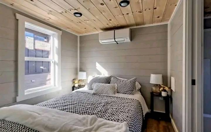 Cozy bedroom with wooden ceiling and gray walls. A bed with patterned linens is centered, flanked by two lamps. Sunlight filters through a window.