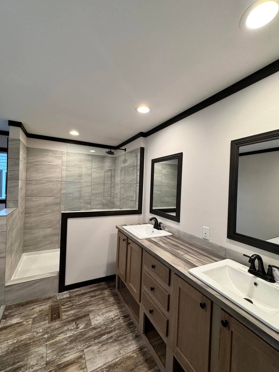 Modern bathroom with dual sinks on a wood vanity, large mirrors, and a glass-enclosed shower. The room has stone tile floors and a serene ambiance.