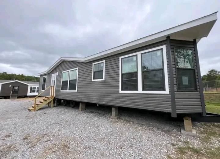 A modern, gray mobile home with large windows sits on concrete blocks on a gravel lot. The sky is overcast, creating a subdued atmosphere.