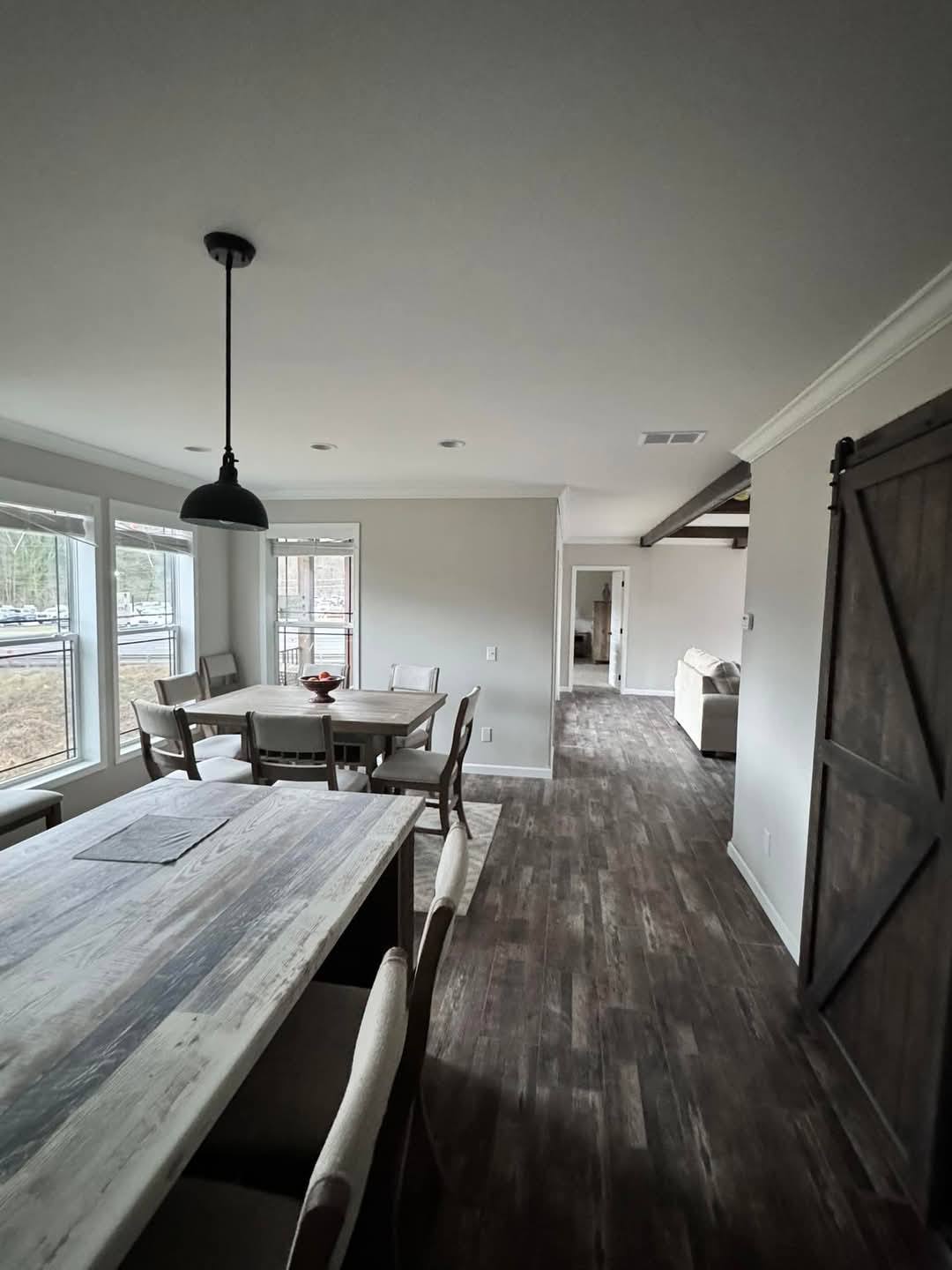 Spacious dining area with a rustic wooden table and chairs, adjacent to a living room. Natural light streams through large windows, creating an inviting, cozy atmosphere.
