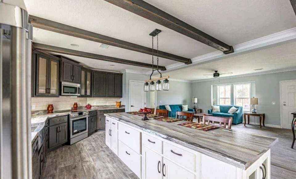 Modern kitchen and living area with wooden beams, a large island, dark cabinets, and stainless steel appliances. Bright, cozy atmosphere with teal sofas.