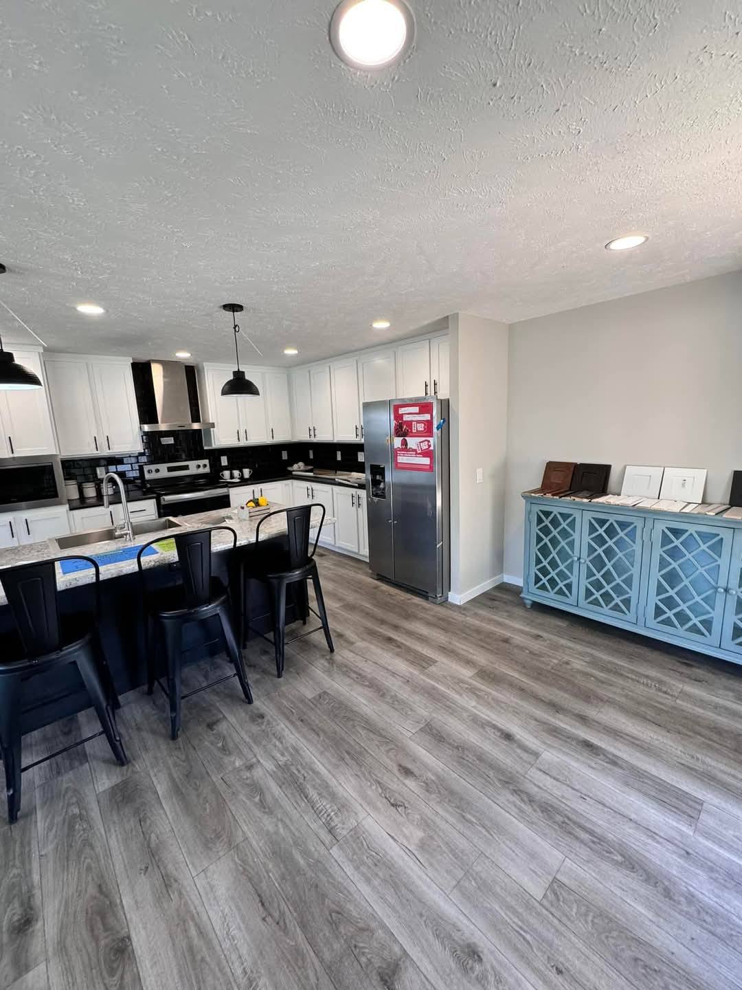 Modern kitchen with white cabinets, black countertops, and stainless steel appliances. An island has black stools and pendant lights. Light wood floors and a blue sideboard add a cozy touch.