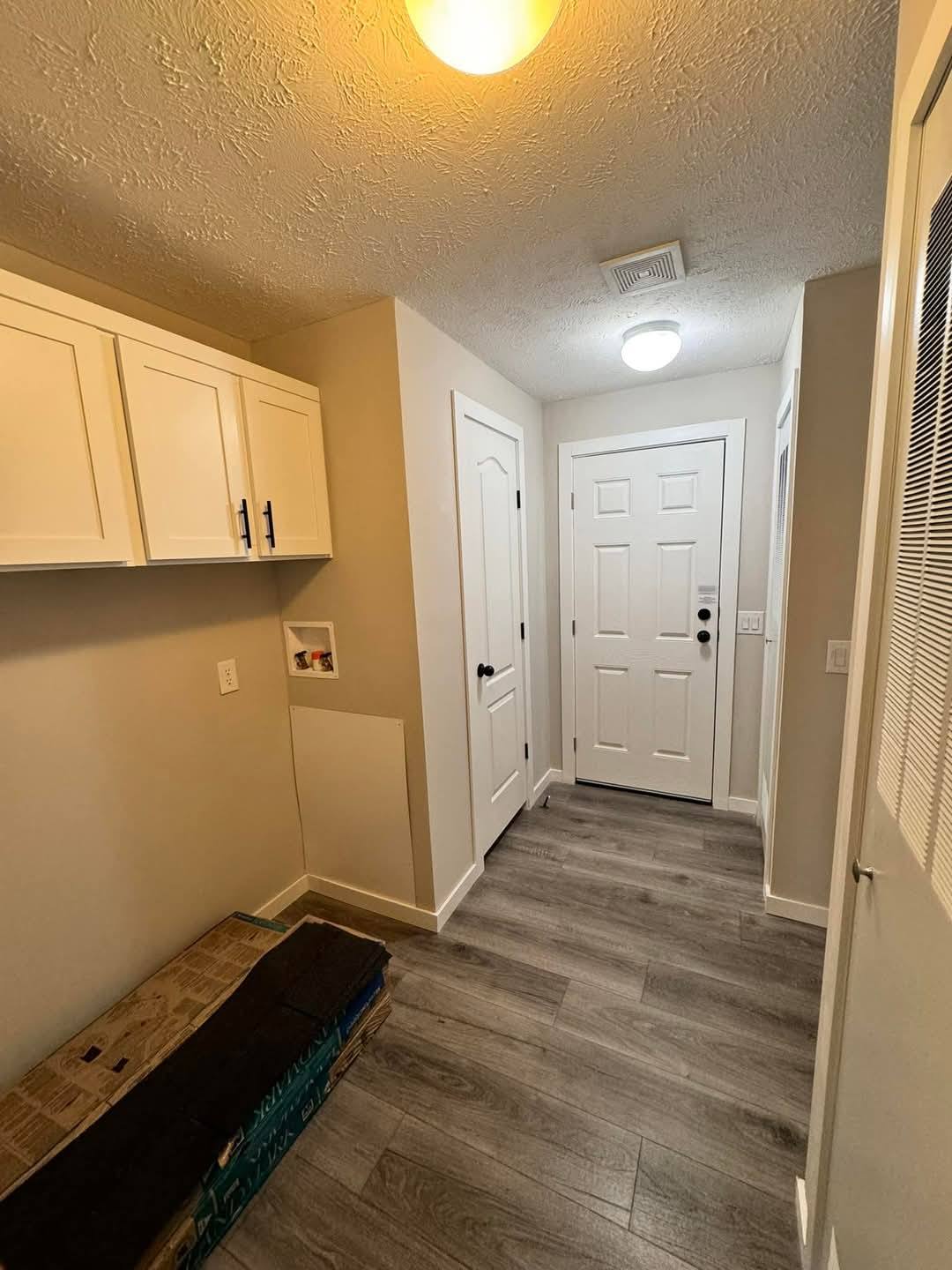 A small, tidy hallway with gray wood floors, white walls, and ceiling lights. Features white cabinets on the left and a closed white door at the end.