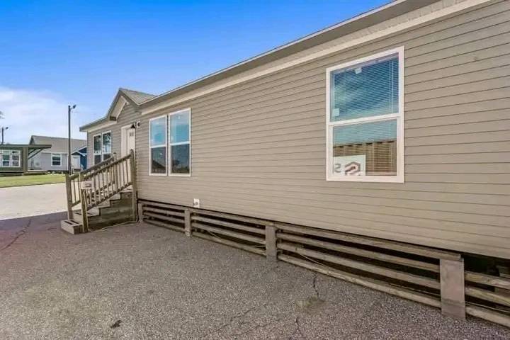 Single-story beige modular home with wooden stairs leading to a small porch. Bright, clear sky above, with another similar home in the background.