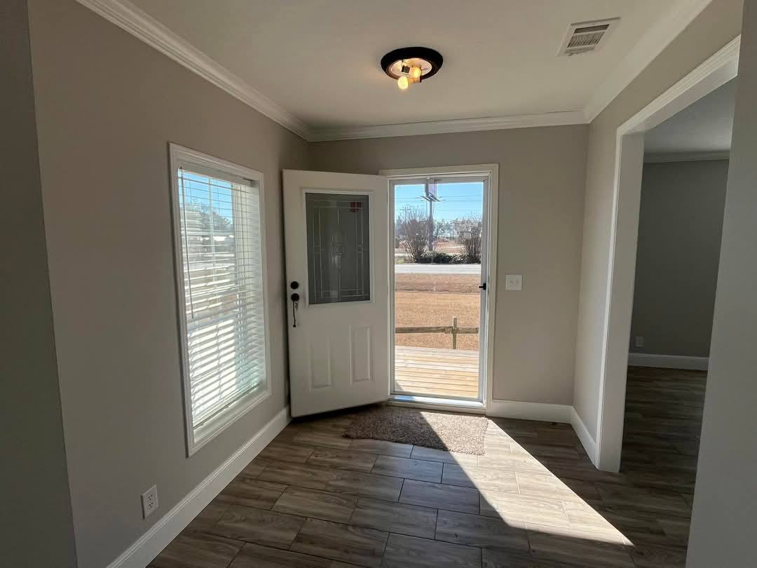 Bright entryway with open door, gray walls, and hardwood floors. Sunlight streams through a glass storm door, casting shadows. Calm and inviting ambiance.