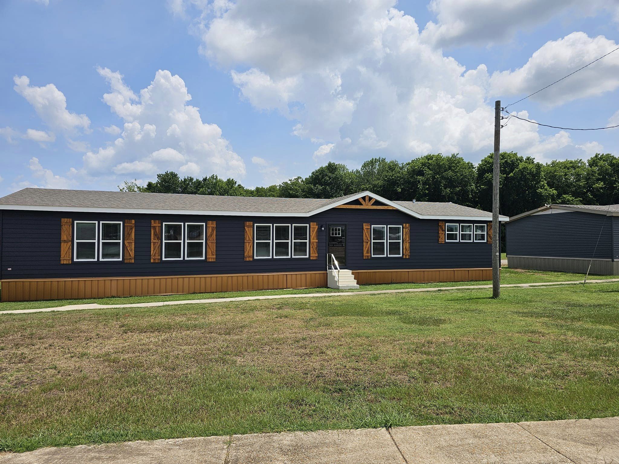 A modern, dark blue manufactured home with white trim and wooden shutters sits under a partly cloudy sky. Lush green trees and grass surround the home.