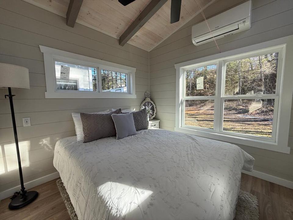 A cozy, well-lit bedroom with wood-paneled walls and ceiling features a large bed with white bedding and gray pillows. Two windows show a sunny forest view.