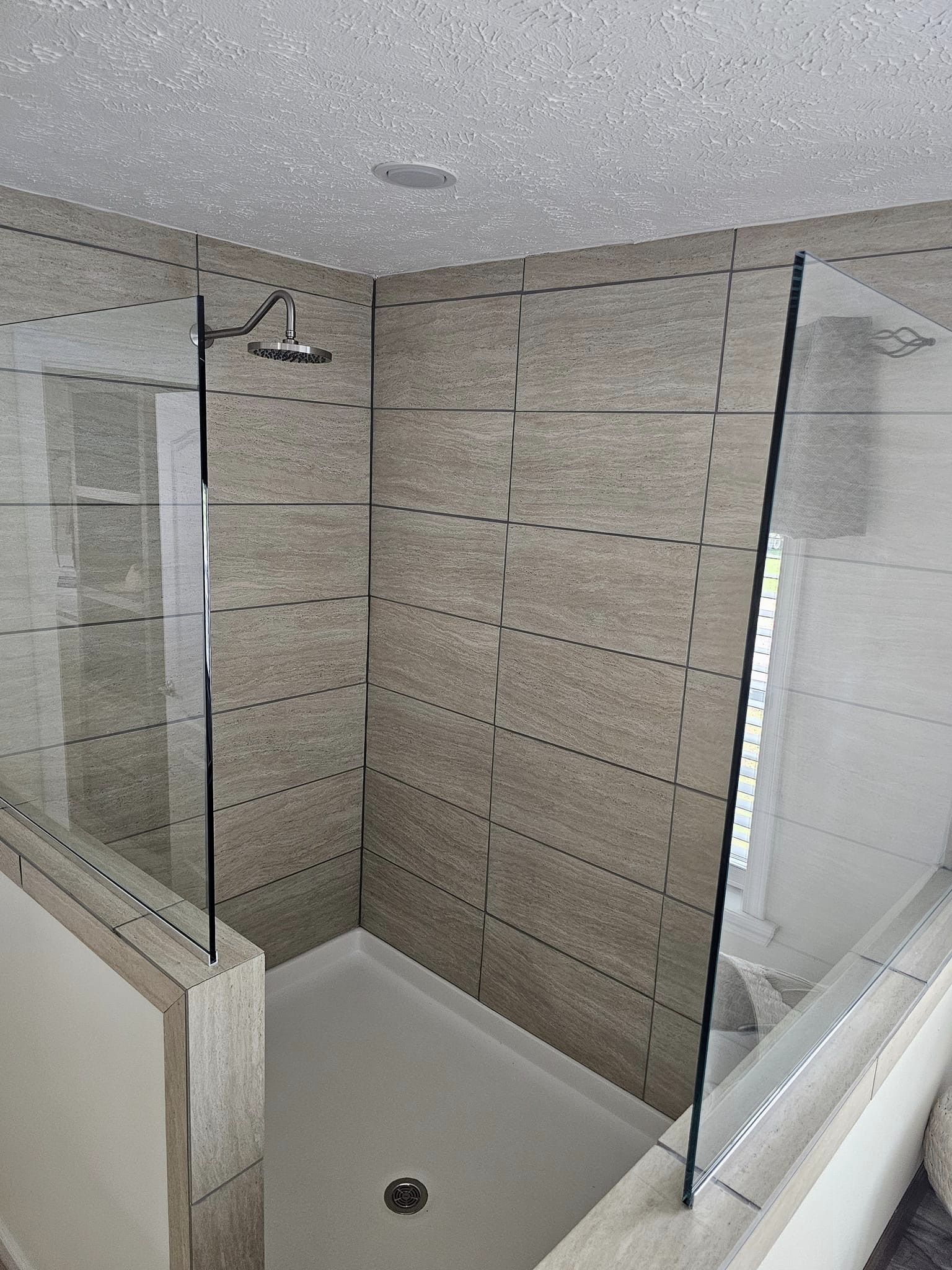 A modern shower with beige tiles and a glass enclosure, featuring a chrome rain showerhead. The space appears clean and well-lit, conveying a sense of freshness and simplicity.